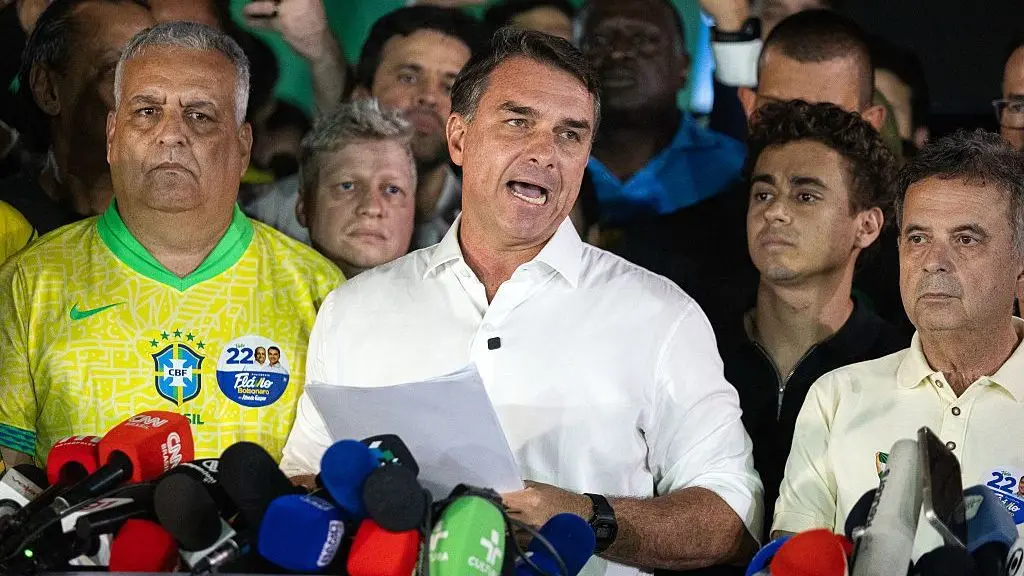 Flávio Bolsonaro viste una camisa blanca manga corta y parece pronunciar un discurso mientras sostiene un papel entre las man